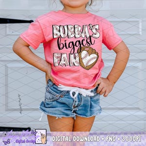 Op de afbeelding: Een roze t-shirt met de tekst "Bubba's Biggest Fan" in zwart en wit, met een honkbalhandschoen met een hartvormige honkbal. Het shirt wordt gedragen met een spijkerbroekshort.