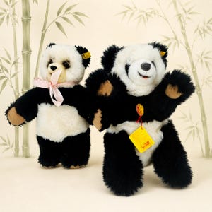 May include: Two plush panda bears with black and white fur. The smaller panda on the left wears a light pink ribbon around its neck. The larger panda on the right features a yellow tag attached to its chest with a red string and a small gold button.