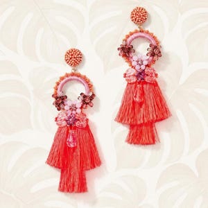Puede incluir: Un par de pendientes de borlas de color coral. Cada pendiente presenta una parte superior con cuentas, un aro rosa y naranja y un dise&ntilde;o de borla en capas. Los pendientes est&aacute;n adornados con detalles florales y un cristal colgante. Fondo blanco con estampado floral.