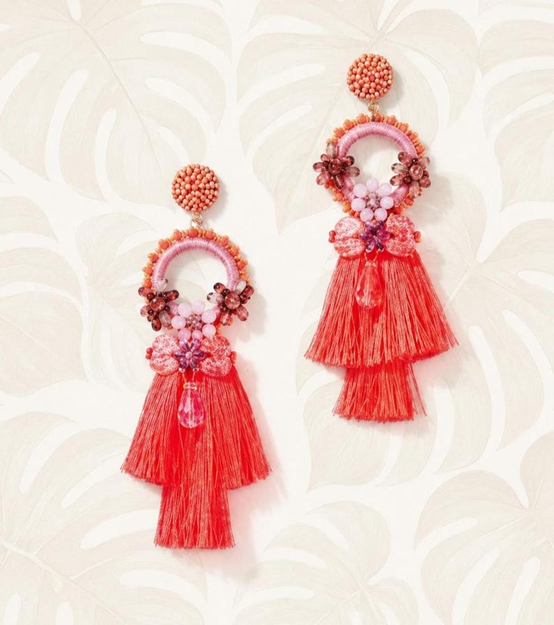 Puede incluir: Un par de pendientes de borlas de color coral. Cada pendiente presenta una parte superior con cuentas, un aro rosa y naranja y un dise&ntilde;o de borla en capas. Los pendientes est&aacute;n adornados con detalles florales y un cristal colgante. Fondo blanco con estampado floral.