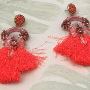 Puede incluir: Un par de pendientes de borlas de color coral. Cada pendiente presenta un peque&ntilde;o bot&oacute;n redondo con cuentas, un aro rosa adornado con peque&ntilde;as cuentas y una borla grande. Los pendientes est&aacute;n sobre un fondo con estampado verde claro y blanco.