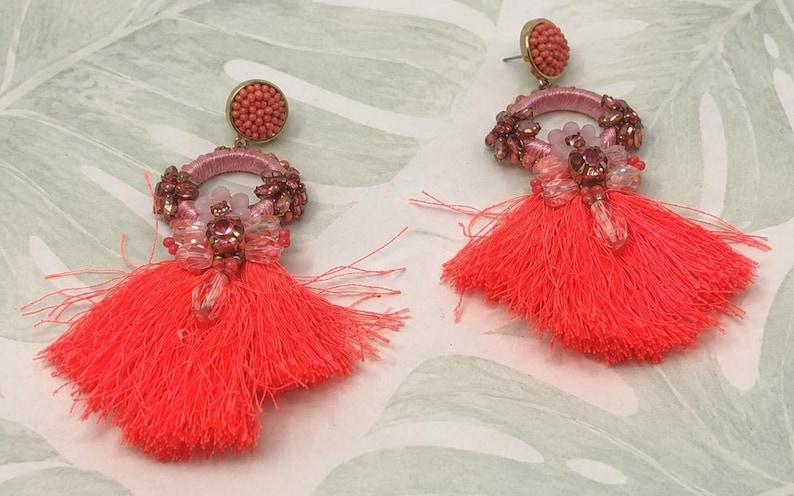 Puede incluir: Un par de pendientes de borlas de color coral. Cada pendiente presenta un peque&ntilde;o bot&oacute;n redondo con cuentas, un aro rosa adornado con peque&ntilde;as cuentas y una borla grande. Los pendientes est&aacute;n sobre un fondo con estampado verde claro y blanco.