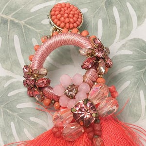 Puede incluir: Primer plano de un pendiente coral y rosa. El pendiente presenta un dise&ntilde;o circular con hilo rosa, cuentas de coral y cristales rosas y transparentes. Una borla coral cuelga de la parte inferior. El pendiente tiene un respaldo dorado.