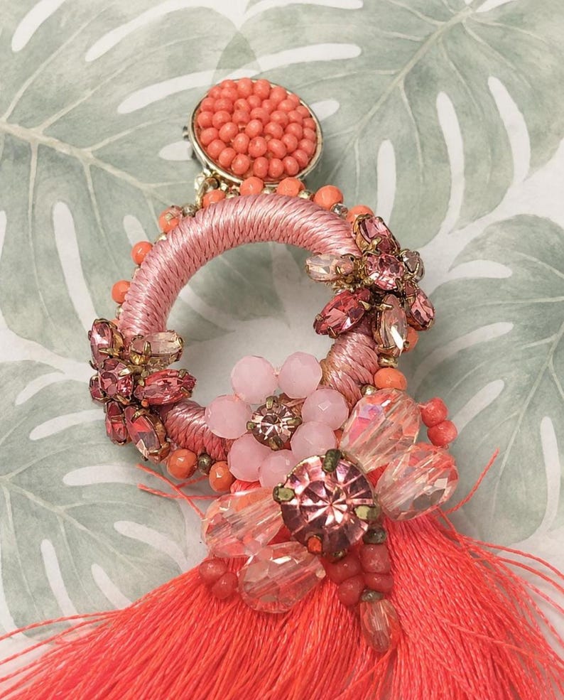 Puede incluir: Primer plano de un pendiente coral y rosa. El pendiente presenta un dise&ntilde;o circular con hilo rosa, cuentas de coral y cristales rosas y transparentes. Una borla coral cuelga de la parte inferior. El pendiente tiene un respaldo dorado.