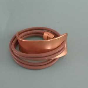 May include: A wrap bracelet featuring a copper-colored metal plate and multiple strands of mauve-colored leather. The bracelet has a magnetic clasp. The design is modern and minimalist.