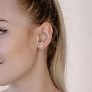 May include: A close-up of a person's ear with a single, round, white pearl stud earring. The earring is centered on the earlobe. The person has blonde hair and fair skin.