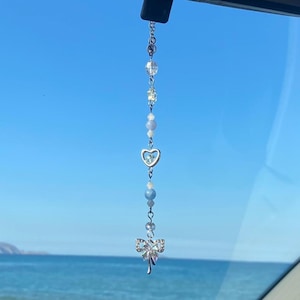 Può includere: Un ciondolo per auto argentato con un design a forma di cuore e fiocco è appeso allo specchietto retrovisore di un'auto. Il ciondolo è in metallo e ha cristalli trasparenti. L'auto è parcheggiata di fronte a un oceano blu.