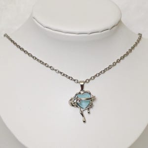 Könnte beinhalten: Halskette mit silberfarbener Kette und Anhänger. Der Anhänger zeigt einen hellblauen, herzförmigen Stein in einem abstrakten Metalldesign. Präsentiert auf einer weißen Büste vor weichem Hintergrund.