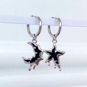 May include: A pair of silver hoop earrings with dangling charms. One charm is a black crescent moon with bat wings and dripping edges. The other charm is a black star with dripping edges.