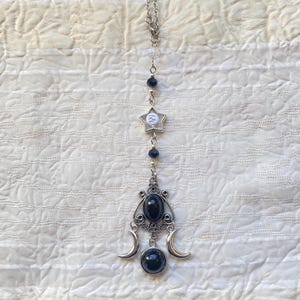 Puede incluir: Un collar de cadena plateada con cuentas negras y un dije plateado con forma de estrella con la letra "W" en el centro. El dije está unido a un dije plateado con dos medias lunas y piedras preciosas negras.