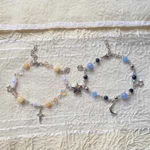 Op de afbeelding: Twee delicate armbanden met ster- en maancharms. De ene armband heeft lichtblauwe en zwarte kralen, terwijl de andere gele en witte kralen heeft.