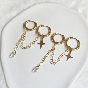 Puede incluir: Cuatro pendientes de aro dorados dispuestos en parejas. Dos aros cuentan con una cadena dorada colgante con una pequeña cuenta transparente, mientras que los otros dos aros presentan un dije en forma de estrella de cuatro puntas.