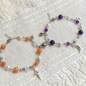 May include: Two silver-toned chain bracelets connected by a magnetic heart charm. One bracelet features orange and white beads with a star charm, while the other displays purple and white beads with a crescent moon charm and a spiral metal accent.