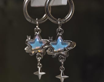 May include: A pair of silver-toned hoop earrings featuring dangling charms. Each charm consists of a central iridescent star surrounded by a thin orbital ring, with a smaller four-pointed star hanging from the bottom of the main star.