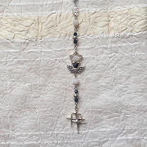 Puede incluir: Un collar de tono plateado con una disposición vertical de cuentas, incluyendo esferas transparentes, blancas y negras. El diseño incorpora un dije en forma de estrella, un par de alas de metal y un colgante de estrella de cuatro puntas.
