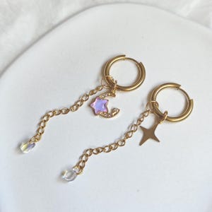 May include: A pair of gold-toned hoop earrings featuring dangling chains. One earring has a crescent moon charm with a purple iridescent star and clear crystals, while the other has a four-pointed star charm. Both chains end in clear beads.