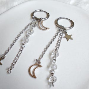 May include: A pair of silver-toned hoop earrings featuring dangling chains, clear faceted beads, and metal charms. One earring displays a crescent moon charm, while the other features both a crescent moon and a five-pointed star charm.