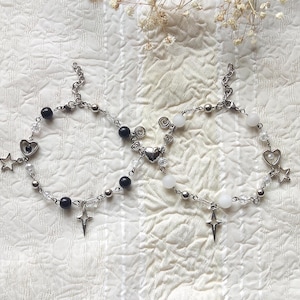 May include: Two silver chain bracelets with black and white beads. One bracelet features a star charm and a heart charm. The other bracelet features a star charm and a heart charm.