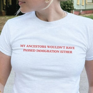 May include: White t-shirt with the red text "MY ANCESTORS WOULDN'T HAVE PASSED IMMIGRATION EITHER." The shirt is short-sleeved and appears to be made of a soft material. The person is wearing gold-colored jewelry and sunglasses.