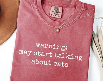 Puede incluir: Camiseta de color carmesí con el texto blanco "warning: may start talking about cats". Un collar plateado con un colgante descansa sobre la camiseta. La camiseta está doblada y tiene una etiqueta "Comfort Colors".