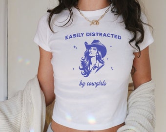 Könnte beinhalten: Weißes, bauchfreies T-Shirt mit einer blauen Grafik einer Cowgirl mit Hut und dem Text "EASILY DISTRACTED by cowgirls".