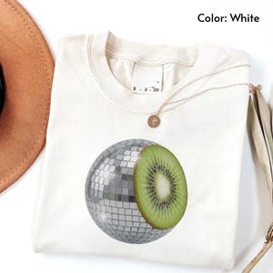 Op de afbeelding: Een wit T-shirt met korte mouwen en een grafische print van een bol die voor de helft uit een zilveren discobal en voor de helft uit een doorgesneden groene kiwi bestaat. Het shirt heeft een ronde hals en een label in de kraag. Tekst: Color: White.