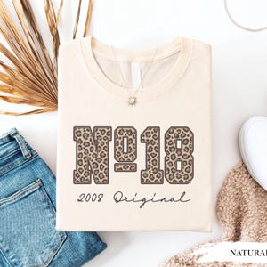 Peut inclure: Un t-shirt couleur crème arborant un grand motif avec le texte "N°18" dans un imprimé léopard marron. Sous le nombre, le texte "2008 Original" est imprimé dans une police cursive noire. Le t-shirt possède un col rond et des manches courtes.