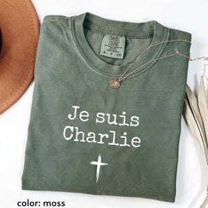 May include: A moss green t-shirt with the text "Je suis Charlie" and a cross graphic in white. A gold-colored necklace with a pendant is draped on the shirt. The shirt is folded and displayed on a white surface.