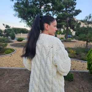 May include: Cream-colored, chunky knit cardigan with a cable-knit design. The long-sleeved sweater is made of thick yarn and has a relaxed fit. The woman is wearing the cardigan outdoors, with trees and greenery in the background.