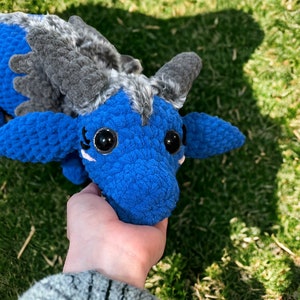 May include: A blue and gray crocheted dragon plush toy with large black eyes. The dragon is being held by a hand.