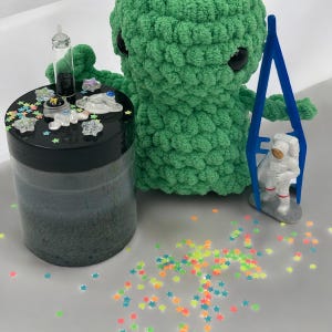 Puede incluir: Una muñeca alienígena verde tejida con ojos negros y un tarro de slime. El tarro tiene una tapa negra con decoraciones temáticas espaciales. Una figura de astronauta está suspendida en un marco azul y confeti en forma de estrella de colores está esparcido.
