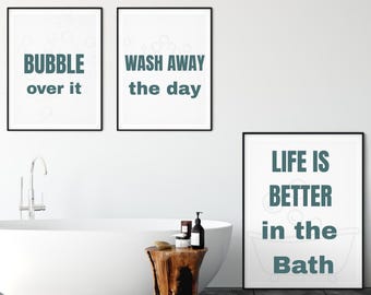 May include: Three framed bathroom art prints with black frames and white backgrounds. The prints have teal text that reads "BUBBLE over it", "WASH AWAY the day", and "LIFE IS BETTER in the BATH". A white bathtub and wooden side table are also in the image.
