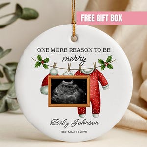May include: A round white ceramic ornament hanging from a gold string. It features the text ONE MORE REASON TO BE merry, a graphic of baby clothes on a clothesline, a framed ultrasound image, and the text Baby Johnson DUE MARCH 202X.