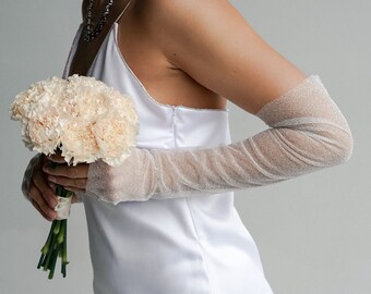 Puede incluir: Un vestido blanco de satén con un diseño sencillo y un ramo de claveles rosa pálido. El vestido tiene tirantes finos y guantes blancos transparentes.