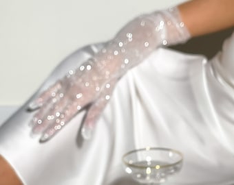 Puede incluir: Guantes blancos transparentes hasta la muñeca con un delicado patrón de pequeñas lentejuelas circulares reflectantes. Los guantes están confeccionados con un tejido de malla ligero y translúcido que permite ver la piel a través del material.