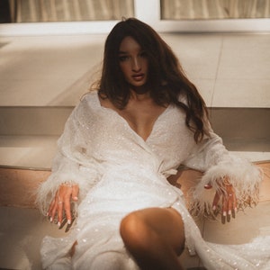 May include: A white, sequined wrap dress with long sleeves and feather trim on the cuffs. The dress has a deep V-neck and is worn by a person sitting on steps. The overall aesthetic is glamorous and elegant.