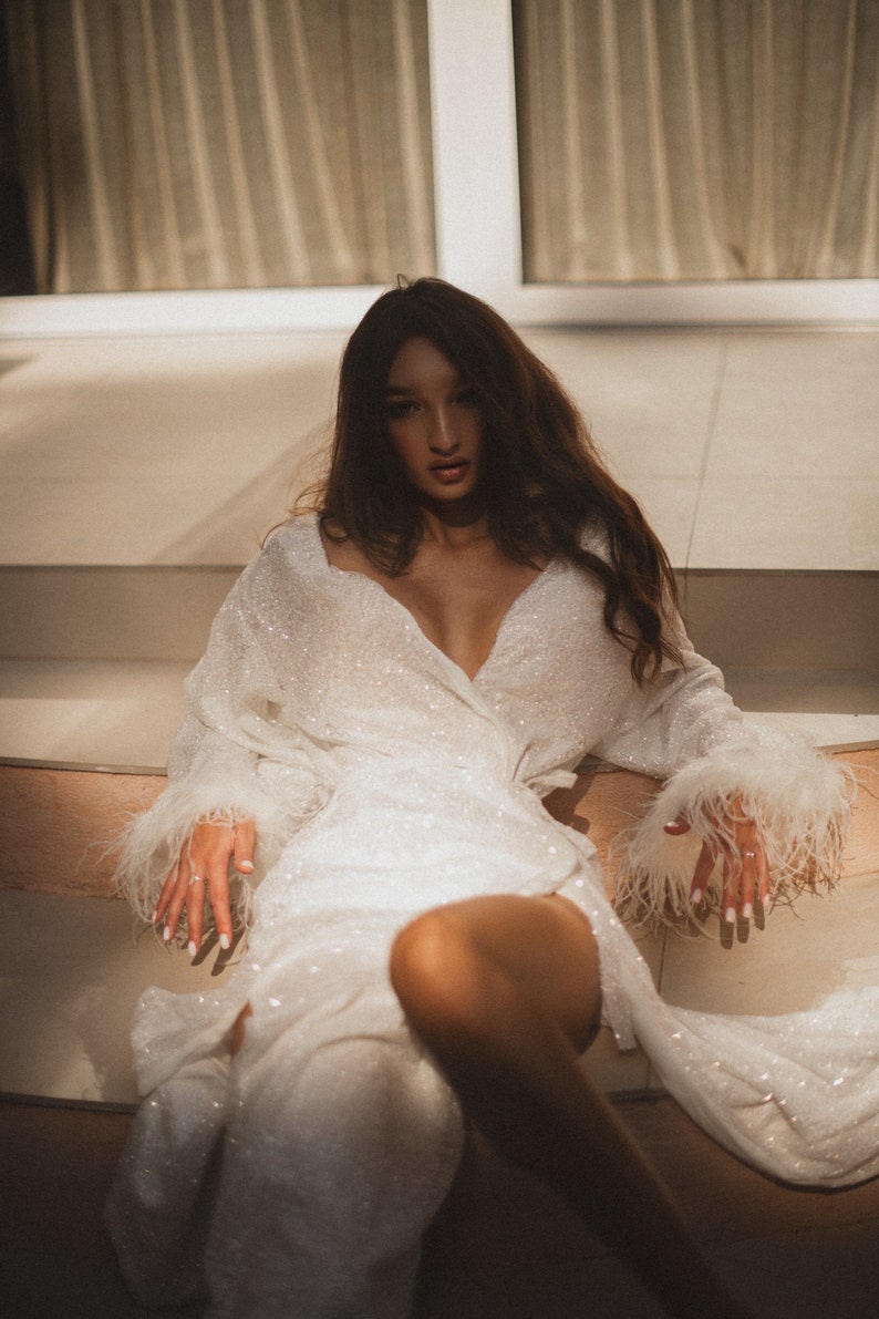 May include: A white, sequined wrap dress with long sleeves and feather trim on the cuffs. The dress has a deep V-neck and is worn by a person sitting on steps. The overall aesthetic is glamorous and elegant.