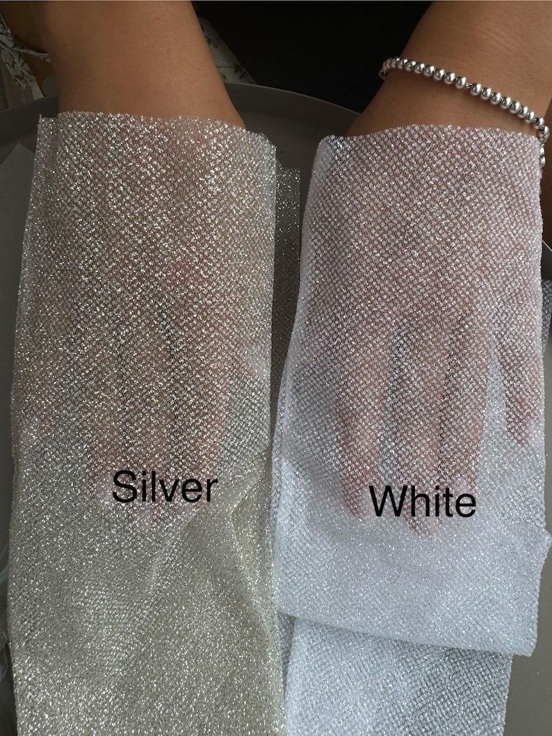 May include: Two pieces of sheer, fine-mesh fabric with a metallic, glittery finish. One piece is labeled Silver and has a warm, reflective tone, while the other is labeled White and features a bright, cool-toned, shimmering appearance.