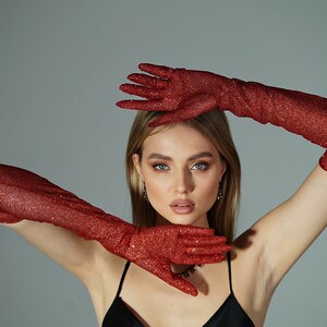 May include: A woman wearing a black sleeveless top and long red sparkly gloves. The gloves are covering her hands and forearms.