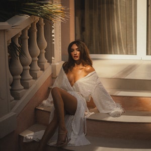 May include: A white, sequined robe with long sleeves and feather trim, worn open, sits on a person on a staircase. The robe is paired with high-heeled sandals. The setting is outdoors, with sunlight and a palm frond visible.