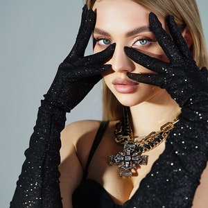 May include: A woman wearing black, sparkly long gloves and a gold chain necklace with a large, jeweled cross pendant.
