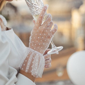 Puede incluir: Una mano de mujer con guantes de encaje blanco con lunares sostiene una copa de cristal de champán.