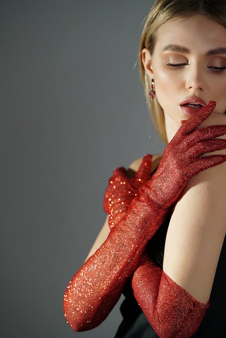 May include: A pair of long red sparkly gloves. The gloves are made of a sheer fabric with a glittery finish. The gloves are worn on the hands of a person with their arms crossed.