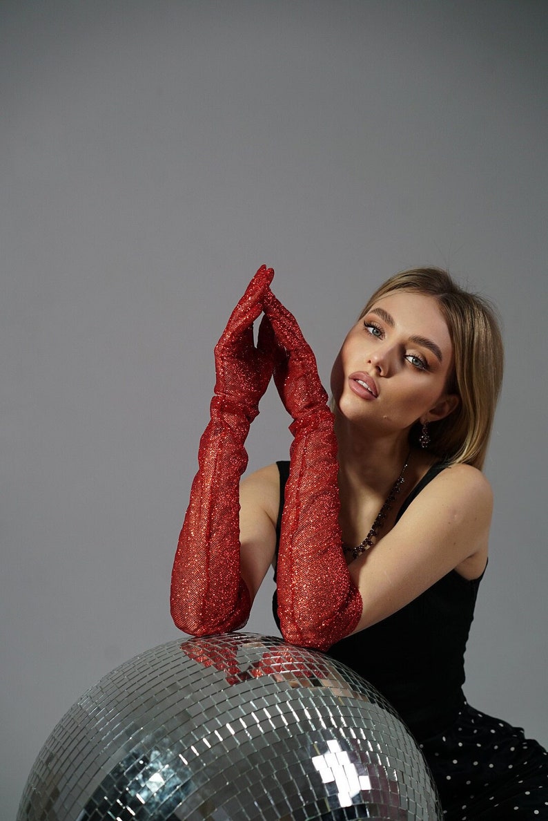 May include: A woman wearing red sparkly gloves poses with a silver disco ball. She is wearing a black tank top and a black and white polka dot skirt.