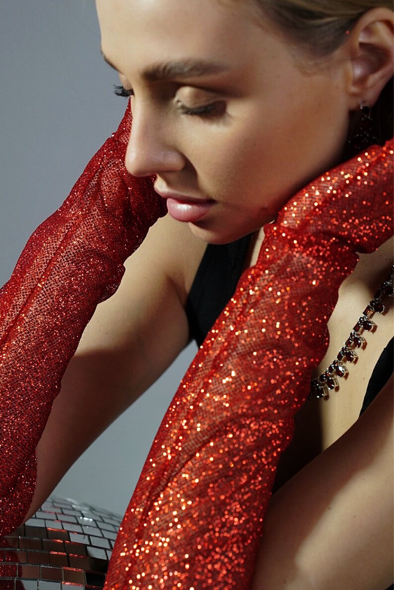 May include: A woman wearing long red sparkly gloves. The gloves are made of a sheer fabric with red glitter embedded in the fabric. The woman is wearing a black top and a necklace with red stones.