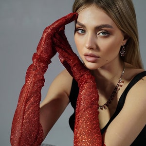 May include: A woman wearing a black sleeveless top and long red sparkly gloves. She is sitting with her elbows resting on a silver disco ball.