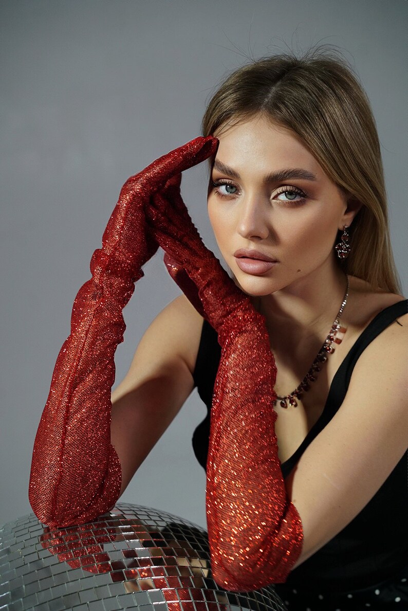 May include: A woman wearing a black sleeveless top and long red sparkly gloves. She is sitting with her elbows resting on a silver disco ball.
