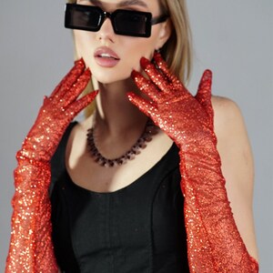 May include: A woman wearing a black top and red glitter long gloves. She is wearing black square sunglasses and a beaded necklace.