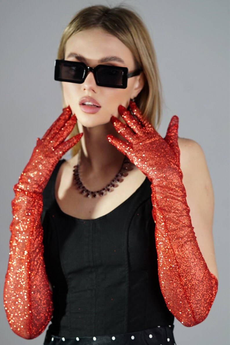 May include: A woman wearing a black top and red glitter long gloves. She is wearing black square sunglasses and a beaded necklace.