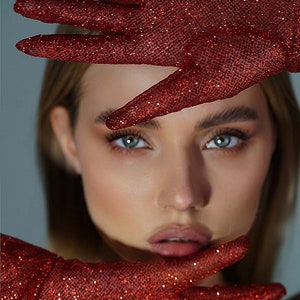 May include: A woman's face is partially obscured by a pair of red sparkly gloves. The gloves are held up to her face, covering her eyes and forehead. The gloves are made of a fine mesh fabric with a red glitter finish.